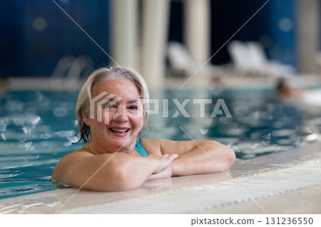 Smiling senior woman enjoying relaxation in an indoor swimming pool, leaning on the poolside. Concept of active lifestyle, wellness, health, hydrotherapy, and leisure for older adults. Smiling senior woman enjoying relaxation in an indoor swimming pool, leaning on the poolside. Concept of active lifestyle, wellness, health, hydrotherapy, and leisure for older adults. 131236550