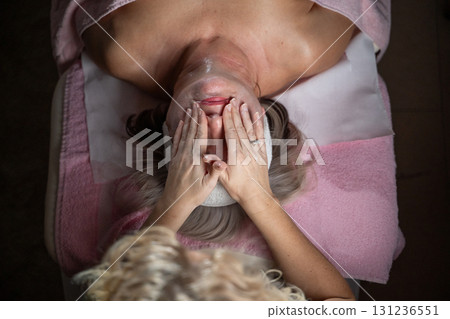 Close up of a mature woman lying on a treatment bed with a towel and headband while a professional therapist applies foam during a facial cleansing procedure. Skincare and wellness concept Close up of a mature woman lying on a treatment bed with a towel and headband while a professional therapist applies foam during a facial cleansing procedure. Skincare and wellness concept 131236551