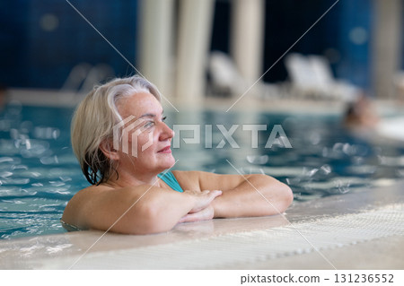 Smiling senior woman enjoying relaxation in an indoor swimming pool, leaning on the poolside. Concept of active lifestyle, wellness, health, hydrotherapy, and leisure for older adults. 131236552