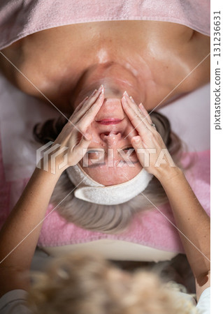 Close up of a mature woman lying on a treatment bed with a towel and headband while a professional therapist applies foam during a facial cleansing procedure. Skincare and wellness concept 131236631