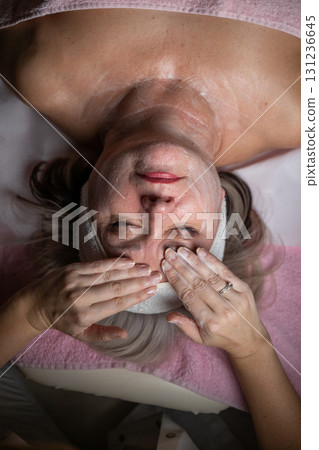 Close up of a mature woman lying on a treatment bed with a towel and headband while a professional therapist applies foam during a facial cleansing procedure. Skincare and wellness concept Close up of a mature woman lying on a treatment bed with a towel and headband while a professional therapist applies foam during a facial cleansing procedure. Skincare and wellness concept 131236645