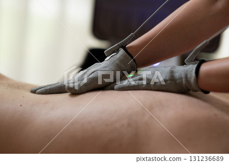 Close up view of a therapist applying electrotherapy with special conductive gloves on a man back during a physiotherapy session. Concept of modern rehabilitation, pain relief, muscle stimulation, and 131236689