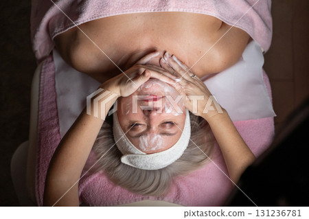 Close up of a mature woman lying on a treatment bed with a towel and headband while a professional therapist applies foam during a facial cleansing procedure. Skincare and wellness concept Close up of a mature woman lying on a treatment bed with a towel and headband while a professional therapist applies foam during a facial cleansing procedure. Skincare and wellness concept 131236781