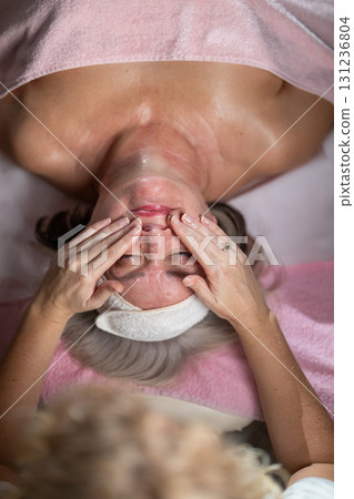 Close up of a mature woman lying on a treatment bed with a towel and headband while a professional therapist applies foam during a facial cleansing procedure. Skincare and wellness concept Close up of a mature woman lying on a treatment bed with a towel and headband while a professional therapist applies foam during a facial cleansing procedure. Skincare and wellness concept 131236804