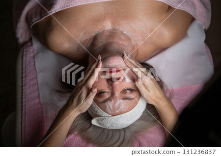Close up of a mature woman lying on a treatment bed with a towel and headband while a professional therapist applies foam during a facial cleansing procedure. Skincare and wellness concept Close up of a mature woman lying on a treatment bed with a towel and headband while a professional therapist applies foam during a facial cleansing procedure. Skincare and wellness concept 131236837
