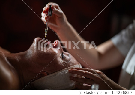 Close up of a mature woman lying in a spa while a therapist applies skincare serum with a dropper above her face. Beauty and wellness concept highlighting anti aging treatment, hydration, and holistic Close up of a mature woman lying in a spa while a therapist applies skincare serum with a dropper above her face. Beauty and wellness concept highlighting anti aging treatment, hydration, and holistic 131236839