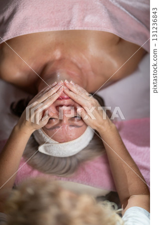 Close up of a mature woman lying on a treatment bed with a towel and headband while a professional therapist applies foam during a facial cleansing procedure. Skincare and wellness concept Close up of a mature woman lying on a treatment bed with a towel and headband while a professional therapist applies foam during a facial cleansing procedure. Skincare and wellness concept 131236843