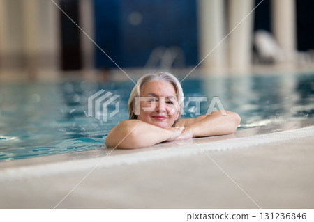 Smiling senior woman enjoying relaxation in an indoor swimming pool, leaning on the poolside. Concept of active lifestyle, wellness, health, hydrotherapy, and leisure for older adults. 131236846