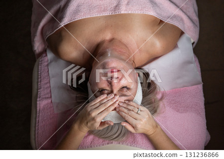 Close up of a mature woman lying on a treatment bed with a towel and headband while a professional therapist applies foam during a facial cleansing procedure. Skincare and wellness concept 131236866
