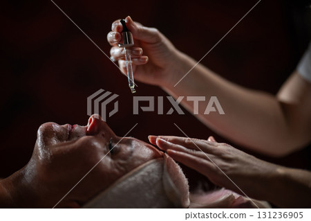 Close up of a mature woman lying in a spa while a therapist applies skincare serum with a dropper above her face. Beauty and wellness concept highlighting anti aging treatment, hydration, and holistic 131236905