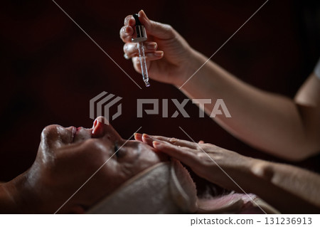 Close up of a mature woman lying in a spa while a therapist applies skincare serum with a dropper above her face. Beauty and wellness concept highlighting anti aging treatment, hydration, and holistic Close up of a mature woman lying in a spa while a therapist applies skincare serum with a dropper above her face. Beauty and wellness concept highlighting anti aging treatment, hydration, and holistic 131236913