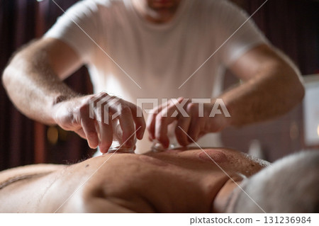 Close up of therapist applying cupping therapy on male clients back during wellness and rehabilitation treatment, traditional alternative medicine for relaxation and pain relief. 131236984