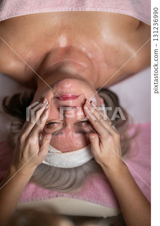 Close up of a mature woman lying on a treatment bed with a towel and headband while a professional therapist applies foam during a facial cleansing procedure. Skincare and wellness concept 131236990