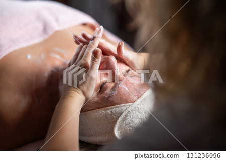 Close up of a mature woman lying on a treatment bed with a towel and headband while a professional therapist applies foam during a facial cleansing procedure. Skincare and wellness concept 131236996