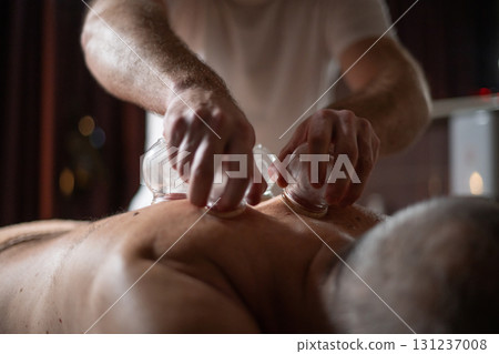 Close up of therapist applying cupping therapy on male clients back during wellness and rehabilitation treatment, traditional alternative medicine for relaxation and pain relief. Close up of therapist applying cupping therapy on male clients back during wellness and rehabilitation treatment, traditional alternative medicine for relaxation and pain relief. 131237008