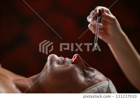 Close up of a mature woman lying in a spa while a therapist applies skincare serum with a dropper above her face. Beauty and wellness concept highlighting anti aging treatment, hydration, and holistic 131237010
