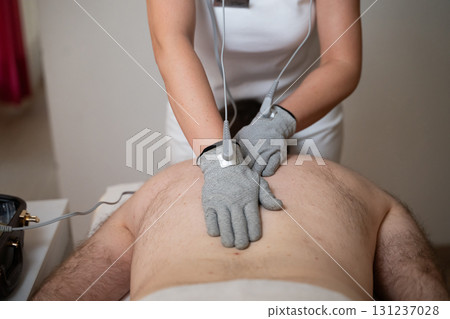 Close up view of a therapist applying electrotherapy with special conductive gloves on a man back during a physiotherapy session. Concept of modern rehabilitation, pain relief, muscle stimulation, and 131237028