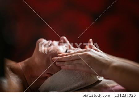 Side view close up of a mature woman lying on a treatment bed with a headband while a professional therapist massages her face. Side view close up of a mature woman lying on a treatment bed with a headband while a professional therapist massages her face. 131237029