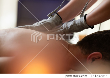 Close up view of a therapist applying electrotherapy with special conductive gloves on a man back during a physiotherapy session. Concept of modern rehabilitation, pain relief, muscle stimulation, and Close up view of a therapist applying electrotherapy with special conductive gloves on a man back during a physiotherapy session. Concept of modern rehabilitation, pain relief, muscle stimulation, and 131237056