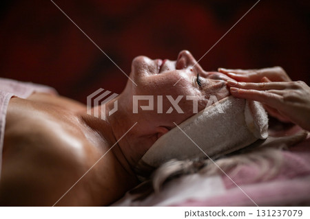 Side view close up of a mature woman lying on a treatment bed with a headband while a professional therapist massages her face. Side view close up of a mature woman lying on a treatment bed with a headband while a professional therapist massages her face. 131237079