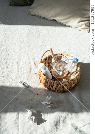 Medication basket with pills, glass of water, thermometer and used tissues on white bed. Medication basket with pills, glass of water, thermometer and used tissues on white bed. 131237995
