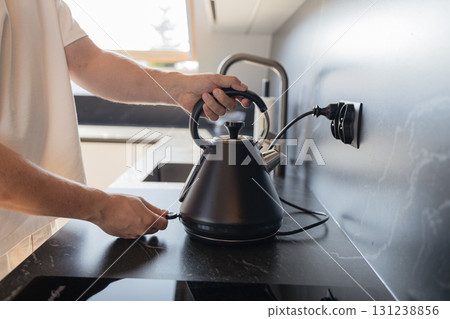 Man placing black electric kettle on modern kitchen stove 131238856
