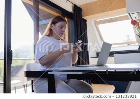 Woman using smartphone while working on laptop at home 131238921