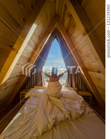 Woman reaching to part curtains at an A-frame cabin window while wrapped in a blanket, revealing a mountain vista outside.  131239360