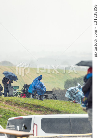 Spectators sit on chairs under heavy rain with umbrellas and rain ponchos, showing resilience and dedication at sport event. Wet weather countryside fans continue follow outdoor sporting tournament 131239859