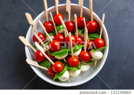 Fresh canape skewers with cherry tomatoes, mozzarella balls and basil leaves served in white bowl. The colorful appetizer reflects Italian caprese style, perfect for party snacks or festive catering 131239860