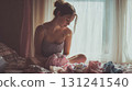 A young woman sits on a bed, tenderly folding small baby clothes in a sunlit room, a moment of peaceful anticipation and care 131241540
