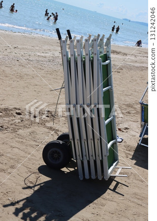 Stack of beach chairs on cart by the sea with people bathing Stack of beach chairs on cart by the sea with people bathing 131242046