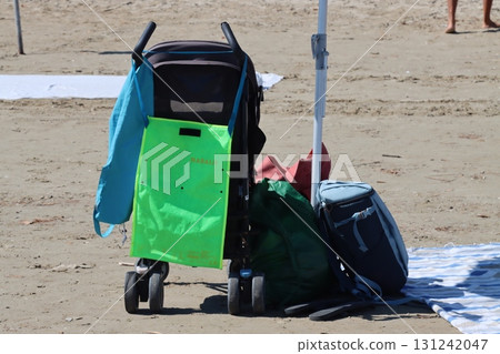 Baby stroller and bags parked on sandy beach under beach umbrella Baby stroller and bags parked on sandy beach under beach umbrella 131242047