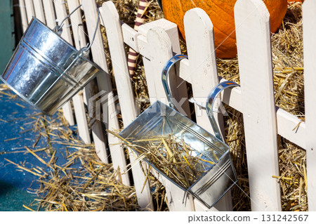 White picket fence adorned with shiny metal containers and straw, crafting a charming autumn harvest display, showcasing a vibrant pumpkin as seasonal decoration White picket fence adorned with shiny metal containers and straw, crafting a charming autumn harvest display, showcasing a vibrant pumpkin as seasonal decoration 131242567