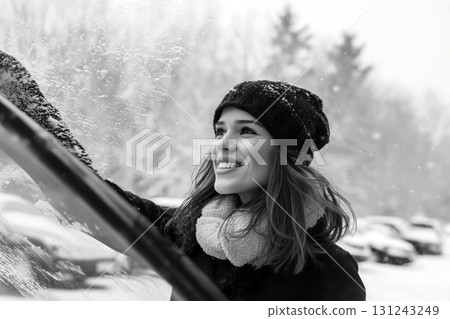 Smiling woman in winter clothes clearing snow from a car windshield on a snowy day in a black and white scene 131243249