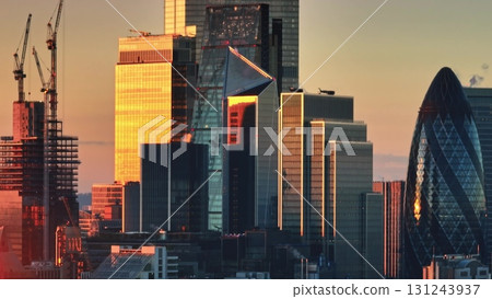 United Kingdom, London: Warm sunset light reflecting off glass facades of London skyscrapers, including The Gherkin, with construction cranes in view. Drone flight 131243937