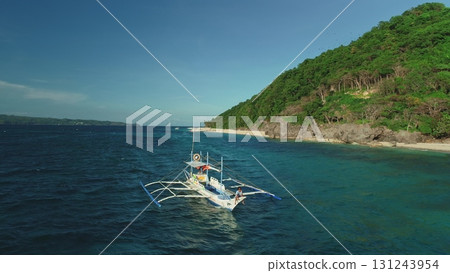 Aerial drone of a traditional bangka boat sailing on turquoise waters near a tropical island with lush vegetation and white sand beach in the Philippines 131243954