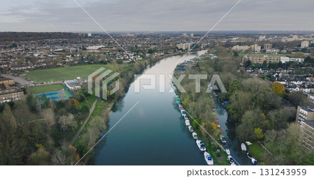 London city , Teddington: River Thames winding through Teddington, showcasing moored boats, residential houses, and green spaces on a cloudy day. UK capital cityscape. Aerial view drone flight 131243959