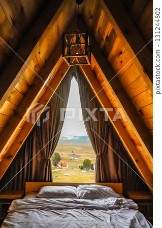 Cozy A-frame wooden cabin interior with a triangular window, white pillows, and blanket. Minimalist rustic design and peaceful retreat atmosphere, perfect for travel and lifestyle imagery. 131244802