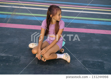 A teenage girl is relaxing while sitting on a skateboard on the asphalt surface of an amusement skate park A teenage girl is relaxing while sitting on a skateboard on the asphalt surface of an amusement skate park 131244998