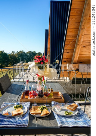 Cozy breakfast on a terrace with toast, eggs, fruits, and wine, overlooking scenic triangular wooden cabins in the mountains of Montenegro. Autumn lifestyle and travel concept. Cozy breakfast on a terrace with toast, eggs, fruits, and wine, overlooking scenic triangular wooden cabins in the mountains of Montenegro. Autumn lifestyle and travel concept. 131245045