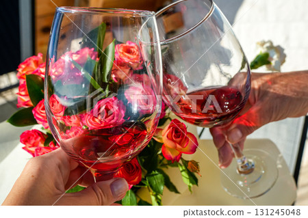 Close-up of two people holding wine glasses by the stem against a floral background. Romantic celebration, lifestyle, and togetherness concept. 131245048