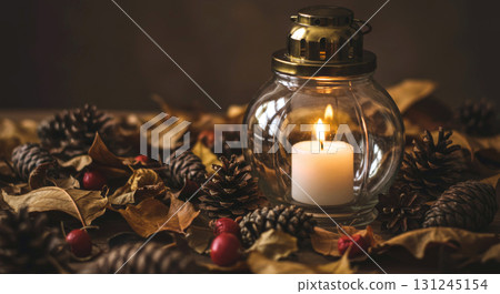 A lit candle in a glass lantern surrounded by pine cones and dried autumn leaves on a wooden surface. 131245154