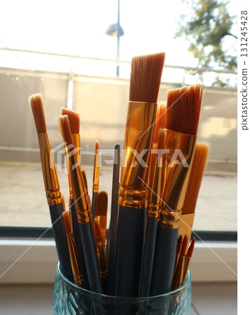 Assorted Gold-Tipped Paintbrushes in Blue Holder Lit by Sunny Window Light for Creative Studio Assorted Gold-Tipped Paintbrushes in Blue Holder Lit by Sunny Window Light for Creative Studio 131245428