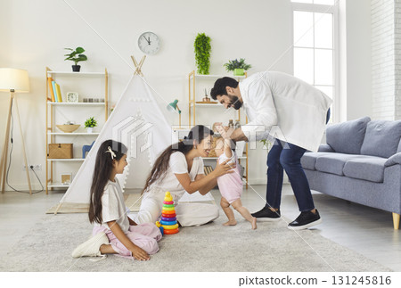 Mother and father teaching toddler to walk with help, happy family playing together in living room 131245816
