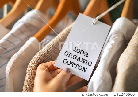 Hand Holding 100 Percent Organic Cotton Label on Eco Friendly Clothes on Wooden Hangers 131246660