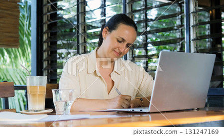 Young woman types on her laptop and writes notes in a notepad on a terrace with tropical greenery while enjoying cold coffee  131247169