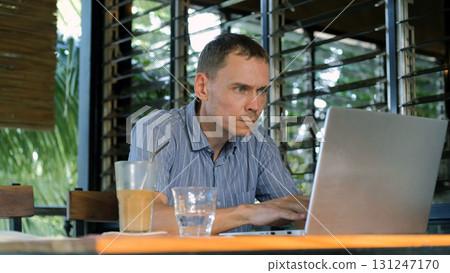 Young man sits on a terrace in a tropical setting focused on a laptop while sipping cold coffee 131247170