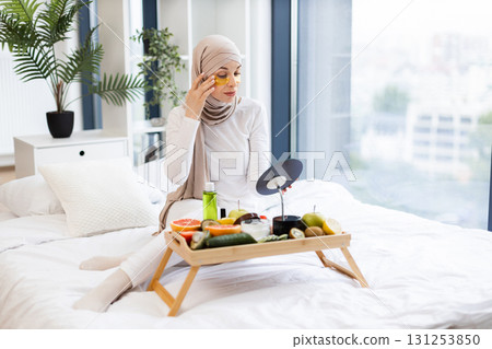 Young Muslim woman in beige hijab on bed handling beauty routine with hydrogel patches, surrounded by fresh fruits, skincare products. Image reflects self-care, health, lifestyle in cozy indoor. 131253850