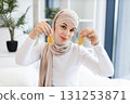 Young Muslim woman focused on skincare routine using gold patches on eye area, showcasing beauty and self-care. Relaxing indoor setting, wearing beige hijab and white top, maintaining wellness. 131253871
